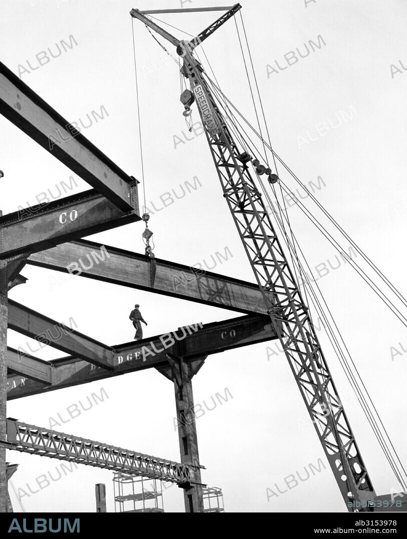 Chicago, Illinois: c. 1938. A high rise steel worker walks a beam while a crane lifts up a new piece of steel.