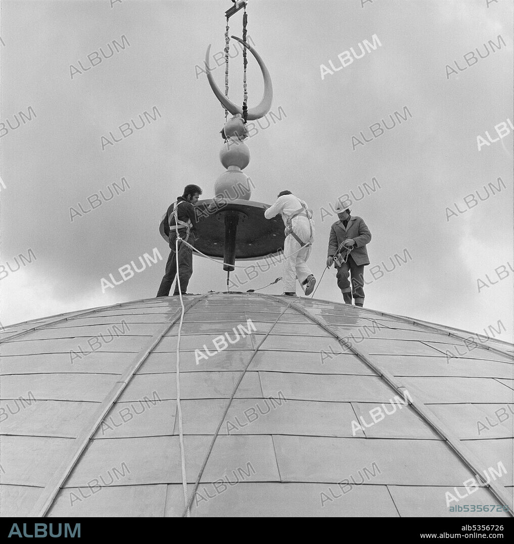 JOHN LAING PLC. London Central Mosque and The Islamic Cultural Centre, Park Road, Regent's Park, City of Westminster, Greater London Authority, 15/03/1977. Men wearing safety harnesses positioning the crescent finial on the domed roof of the prayer hall at the London Central Mosque, as it is lowered from above by a crane. The finial was manufactured from glass-fibre reinforced concrete (GRC) in Northamptonshire, and painted in South Wales to give a gold coloured finish.