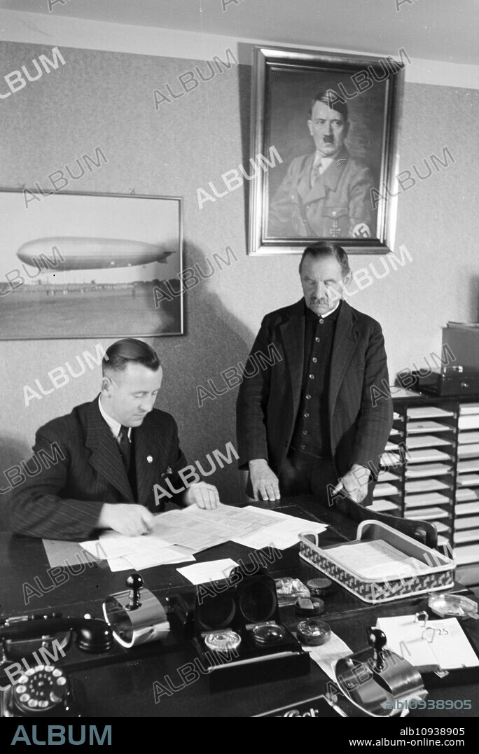 The first mayor of the zeppelin village, Hans von Schiller, and an employee in his bureau with a portrait of Adolf Hitler hanging above the desk, Germany 1930s.