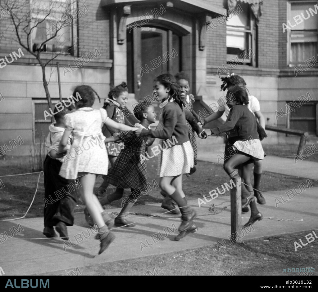 Entitled: "Children playing 'ring around the rosie' in one of the better neighborhoods of the Black Belt, Chicago, Illinois." Ring Around the Rosie is a nursery rhyme or folksong and playground singing game. It first appeared in print in 1881, but it is reported that a version was already being sung to the current tune in the 1790s and similar rhymes are known from across Europe. The first printing of the rhyme was in Kate Greenaway's 1881 edition of Mother Goose; or, the Old Nursery Rhymes: Ring-a-ring-a-roses, A pocket full of posies; Ashes! Ashes! We all fall down. Photographed for the Farm Security Administration by Edwin Rosskam, April 1941.