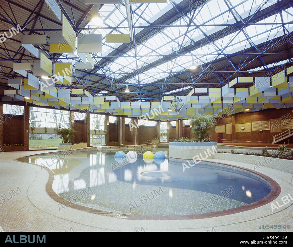JOHN LAING PLC. Kingfisher Leisure Centre, Fairfield Road, Kingston upon Thames, London, 30/11/1983. The beach and lagoon zones of the swimming pool at Kingfisher Leisure Centre, with the acoustic absorbent panels suspended from the roof structure above. In April 1982, a turf-cutting ceremony marked the symbolic start of construction of a new leisure swimming pool, squash courts and cafeteria at Fairfield Road for the Royal Borough of Kingston upon Thames. The scheme design and detailed brief had been provided by the borough in December 1981. The main swimming pool hall was designed to approximate an outdoor pool setting, with a large curvilinear form, shallows accessible by wheelchair, a waterspout, waves and a beach area. A lane zone for competitive swimming and separate teaching pool were also housed in the building. The main pool and beach were lit by natural light through the roof and vertical glazing. Acoustic absorbent panels were suspended from the roof structure; these also contributed to a simulation of outdoor noise dispersal. The completed pool, known as the Kingfisher Leisure Centre, was handed over to the Mayor on 2nd December 1983.