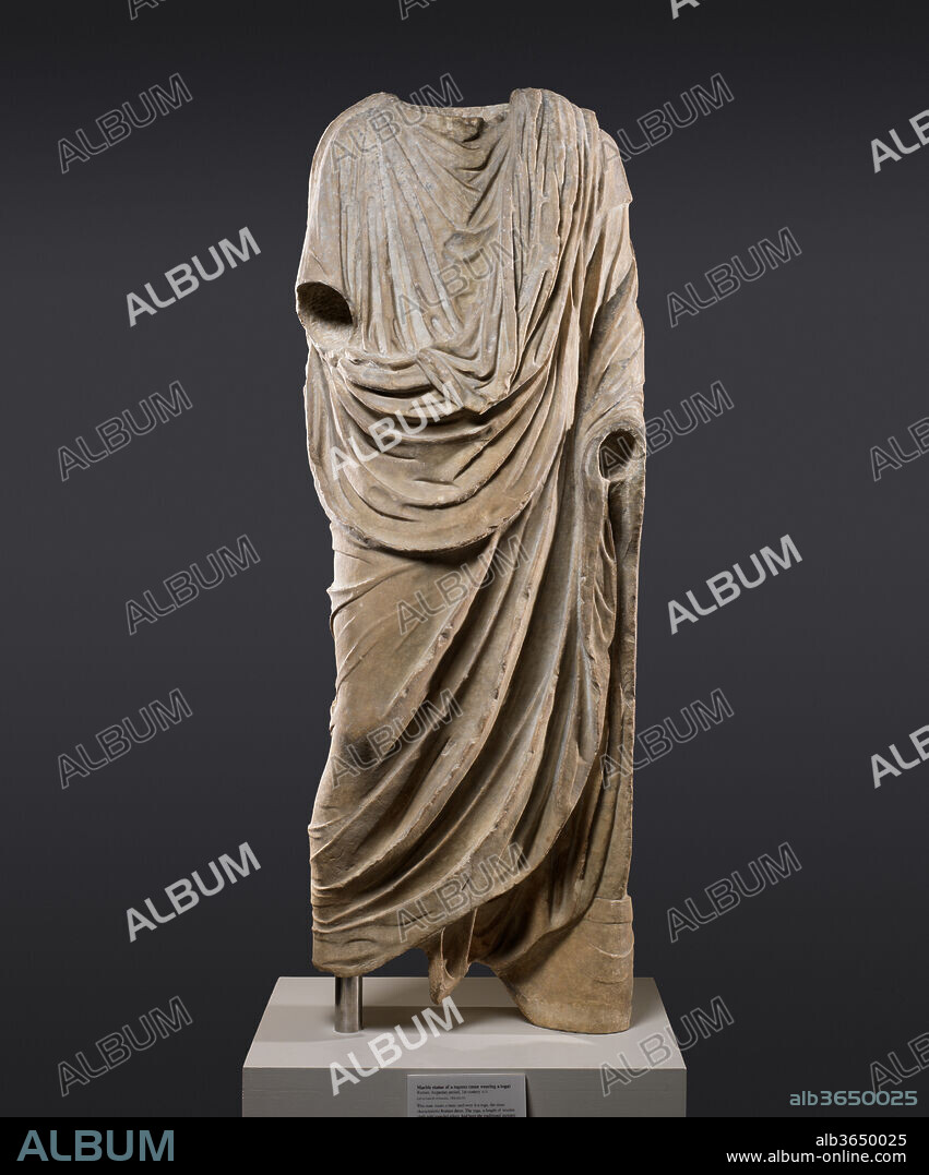 Marble statue of a togatus (man wearing a toga). Culture: Roman. Dimensions: H. 72 in. (182.9 cm). Date: 1st century A.D..
This man wears a tunic and over it a toga, the most characteristic Roman dress. The toga, a length of woolen cloth with rounded edges, had been the traditional garment of the Romans for centuries, but by the late first century B.C., it was declining in popularity. As part of his effort to revive ancient values and customs, the emperor Augustus made the toga a sort of unofficial state dress that all citizens were required to wear in the forum. A cylindrical leather box for scrolls, represented at the feet of this figure, identify him as a man engaged in public business. A portrait head and arms were carved separately and added.