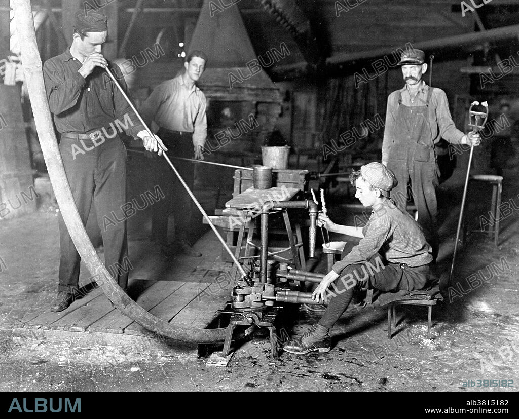 Entitled: "Glass Blower and Mold Boy. Boy has 4 1/2 hours of this at a stretch, then an hour's rest and 4 1/2 more: cramped position. Day shift one week: night shift next. Location: Grafton, West Virginia." There were four main jobs in the glass factories that used boys as a majority of the labor. The boy who opens and closes the iron mold for the blower was the "holding-mold boy". The second job required the boy to stand beside the presser and receive the tumblers from the large mold was called the "ketchin-up boy". The third job was for the boy who seized the blown or pressed objects with a long iron rod and holds them in the flame of the glory hole was called the "sticker-up boy". The final job was for the boy who takes the finished objects from the finisher to the annealing oven for the final firing of the object and was called the "carrying-in boy". These jobs required long hours and cramped conditions. Photographed by Lewis Hine, October 1908.