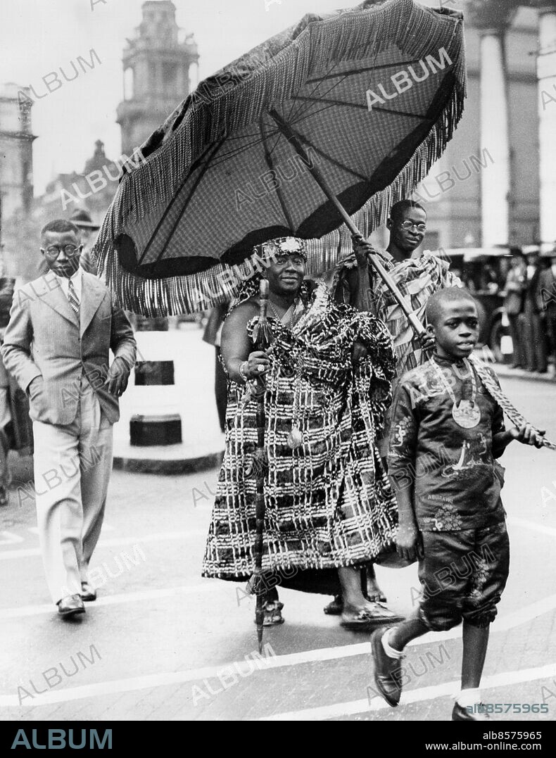 Orig. bildtext... PARAMOUNT CHIEF OF THE GOLD COAST IN LONDON. ACCOMPANIED BY NINE YEAR OLD SWORD BEARER. Accompanied by his nine-year-old sword bearer, who, by tradition, never leaves his side, Sir Ofori Atta, Paramount Chief of the Gold Coast of Africa has arrived in London for a visit. He wears always a gold crown, golden sandals and thick velvet robes. He was considerably interested in the white traffic lines in Trafalgar Square, and insisted on walking across them, preceded by his sword bearer, and covered by his ceremonial umbrella. PHOTO SHOWS:- Sir Ofori Atta crossing the pedestrian lines at Trafalgar Sqaure, London. June 25th 1934 Anm. Nana Sir Ofori Atta I, 1881-1943, kung av Guldkusten 1912-1943. Guldkusten - brittisk koloni 1874-1957 Paraplyer Parasoll Handen i fickan Svärd Svärdbärare Kvartett Kronor Spiror Promenera Kungligheter Traditionell klädsel CD648 persons: NANA SIR OFORI ATTA I, KUNG AV GULDKUSTEN sites: AFRIKA*;GULDKUSTEN*;LONDON;STORBRITANNIEN.
