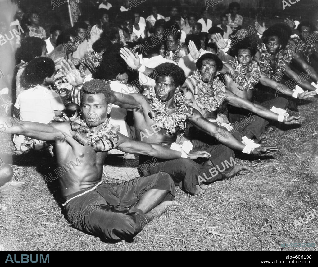 Fiji, Men perform a 'vakamalolo'A group of young Fijian men sit cross-legged in a line and stretch out their arms as they perform a 'vakamalolo' or sitting dance. Naked from the waist up, they wear ceremonial costume including face paint and leis made from leaves. Caption reads: A Vakamalolo which tells a story of some event of the past, 1965. 2005/010/1/14/40.