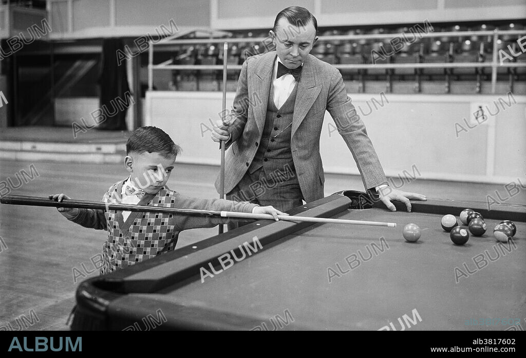 World's champion watches Washington baby cue wizard perform with ivories. Just reaching his sixth birthday and barely able to see over the table, Andrew M. Dargan, son of a Washington pool room owner, is amazing pocket billiard expert with his mastery of intricate shots on the green cloth. He is shown in this photograph taking a few pointers from Irwin Rudolph, world's champion at pocket billiards who is in Washington for an exhibition. Erwin Rudolph (December 30, 1893 - May 19, 1957) was an American pocket billiards player from Cleveland, Ohio and a four-time world champion. One of his great feats was running 125 points in 32 minutes (now eclipsed). was inducted to the Billiard Congress of America Hall of Fame in 1987, Pool, also more formally known as pocket billiards (mostly in North America) or pool billiards (mostly in Europe and Australia), is the family of cue sports and games played on a pool table having six receptacles called pockets along the rails, into which balls are deposited as the main goal of play. Popular versions include eight-ball and nine-ball. An obsolete term for pool is six-pocket. Photographed by Harris & Ewing, 1927.