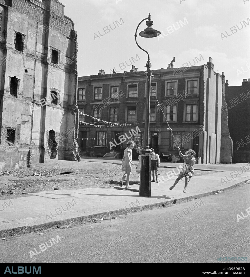Children swinging on a lamppost, London, 1960-1965. Children swinging on ropes tied to a lamppost beside the site of a demolished terraced house in either Islington or Camden. There is bunting across the road between terraced houses behind them.