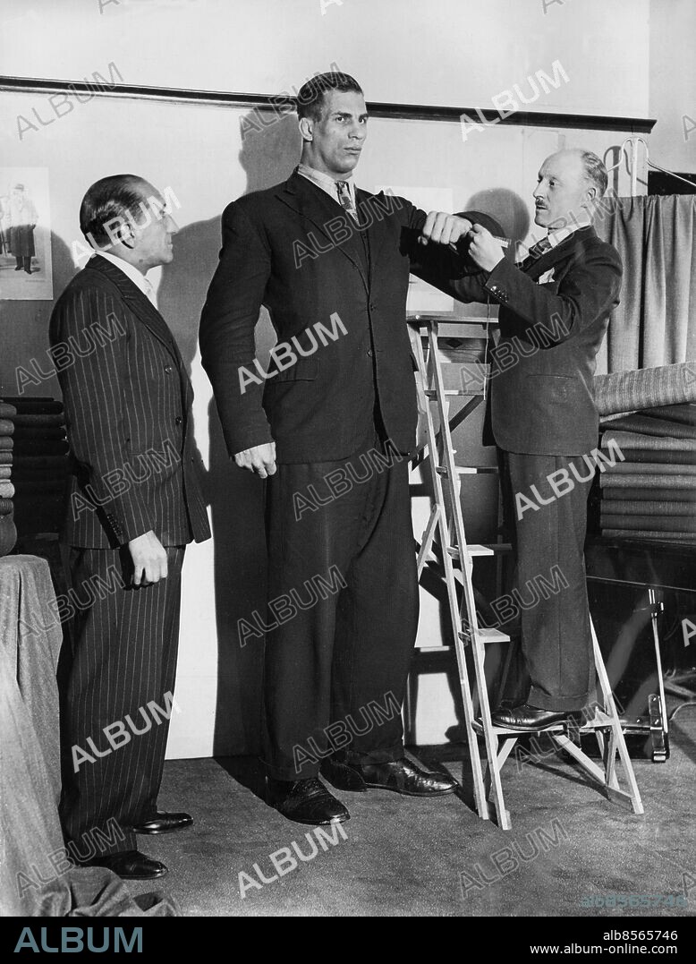 Orig. bildtext... MEASURING NEW MAN MOUNTAIN FOR A SUIT! 7-FOOT POLISH BOXER IN LONDON. Taller even than Primo Carnera, 7 feet 3 inches Leon Ketchell, boxer, wrestler, footballer and swimmer of Warsaw, har arrived in London in search of world boxing honours. Although only 24 years old, Ketchell is 7 inches taller than Carnera and weighs 20 stones. He wears a size 20 collar, takes 18 shoes and has a chest measurement of 48¬? inches. Ketchell is willing to challenge the champions either in boxing or wrestling. It is planned to place him under the care of Larry Gains for tuition. PHOTO SHOWS: Leon Ketchell being measured for a suit in London. On left is Harry Levene, the famous boxing manager. Anm. Mått Mätningar Brottare Stegar Kostymer Tyger Randigt Måttagning Jättar Trio Storvuxna Jämförelser CD308 persons: HARRY LEVENE;LEON KETCHELL sites: POLEN*;STORBRITANNIEN;LONDON.