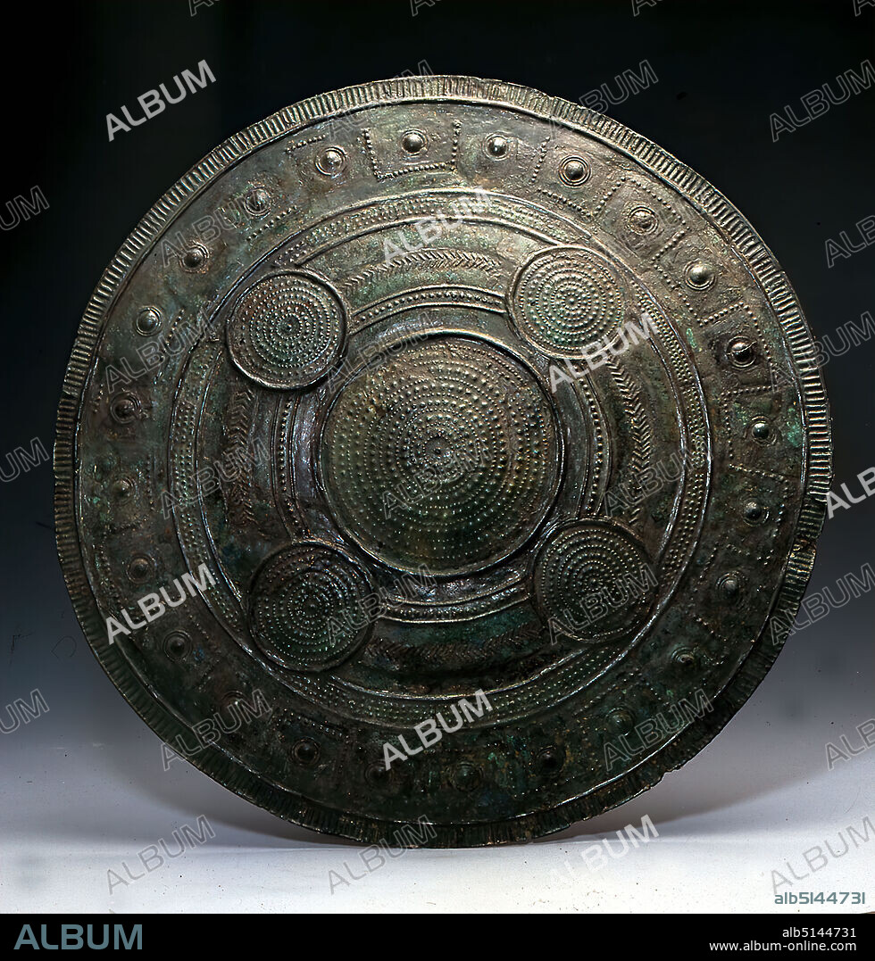 Round Shield, Bronze, chased, Bronze, Total: Diameter: 56.5 cm, Weapons (warfare/military), Armor (warfare/military), Burial objects, Ritual objects and accessories, Antiquity, The flattened shield has a more strongly curved hump in the center. The decoration is chased. On the large shield hump seven rows of dots alternate with six rows of humps. There is a hole in the central circular hump. The same ornamental system is repeated in four circular fields, each of which is surrounded by a strong border strip and arranged in such a way that together with the hump (lat. umbo) they form a five-point arrangement. At the top it cuts through a line of humps running around the entire shield and also framed by border strips, at the bottom they protrude into an identical circular line filled with three rows of smaller humps. Between the four circular fields are angular rows or abstract branches. Towards the edge follows a wide zone of simple meandering, consisting of small humps, in whose individual sections there are large humps surrounded by two circular lines. From this, the border strip is separated by a strip with rays. The earliest bronze shields appear during the late 8th century BC in southern Etruria. They are the typical feature of rich Etruscan chamber tombs and document the transition from the Villanova culture to the Etruscans. The majority of shields from this period were found in warrior tombs with extensive armor.