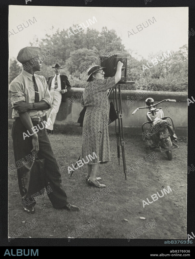 Photographer Frances Benjamin Johnston standing beside her view camera preparing to take a photograph Biltmore Estate; her assistant Huntley Ruff is in the foreground and a motorbike is in the background, 1938.