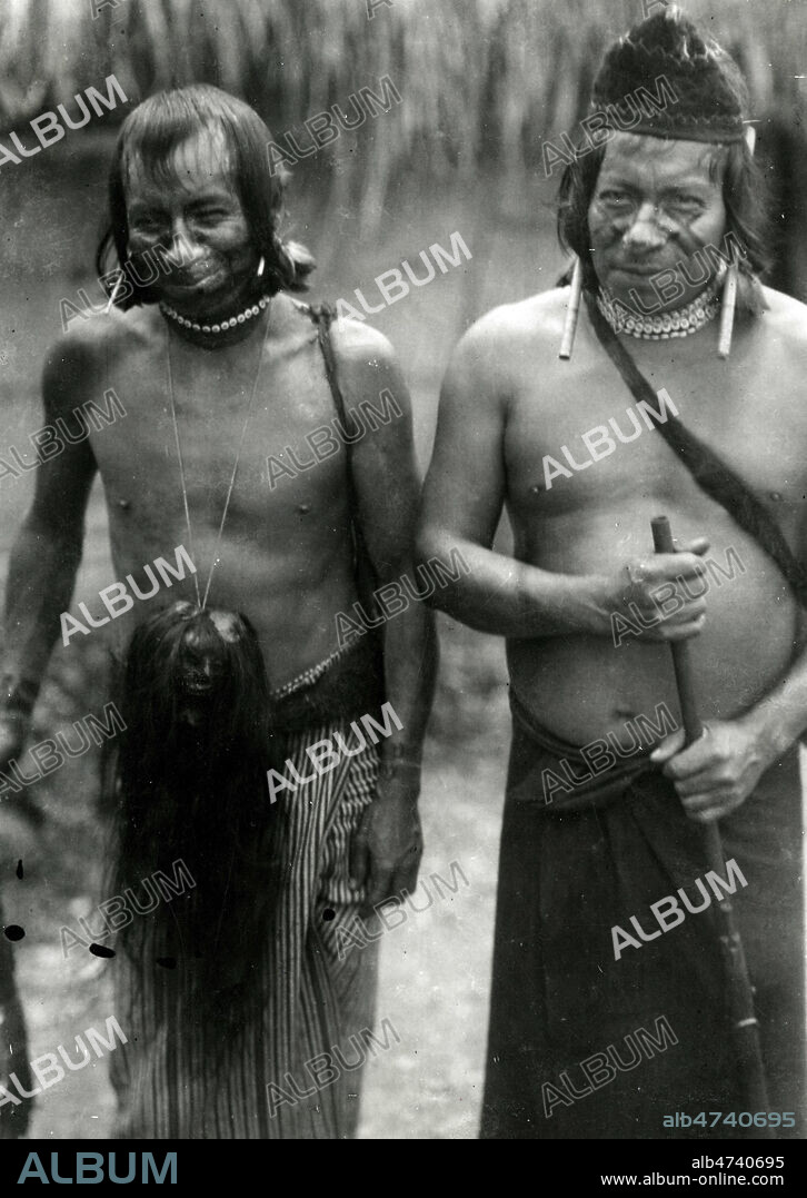 Deux types de Jivaros (aujourd'hui appeles Shuars, ou Aguarunas, peuple amerindien , entre l'Equateur et le Perou) , l'un d'eux porte suspendu en trophee la tete reduite d'un ennemi abattu. Amazonie. Photographie fin 19eme debut 20eme siecle. Collection privee. Credit Collection NBL/KHARBINE TAPABOR.