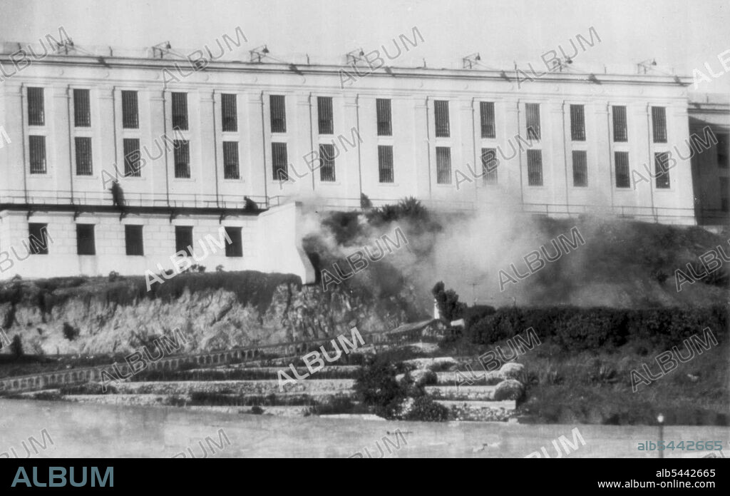 Guards Battle Convicts In Alcatraz Riot -- Guards creep along beneath cellblock windows to fire into building where convicts, armed with machine gun are holding most of Prison officers here late today. Warden James A. Johnston had issued a riot call. Note fire burning in Foreground, which is believed to have started from tracer bullets fired by convicts. May 2, 1946. (Photo by AP Wirephoto).