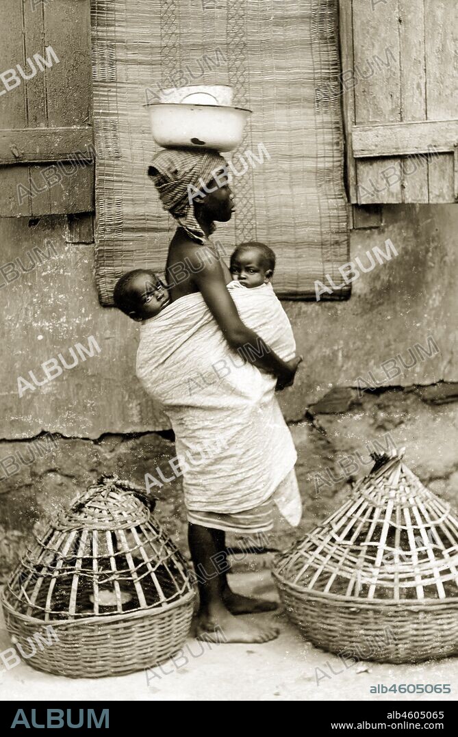 Nigeria, Marking on reverse: 'A young Yoruba girl wife with her two 'pickins''. Previously to the picture she was carrying one basket inside the other on her head besides what she has on her head here. They are baskets for chickens and are very heavy although they do not look it. Note the native mat hanging at the window used in that home as a curtain', [c.1920s]. 2000/098/2/82.