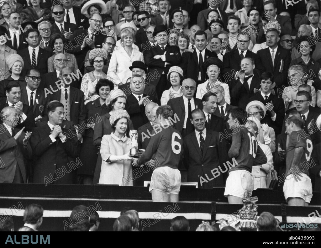 Orig. bildtext... WORLD CUP FINAL - ENGLAND V. WEST GERMANY AT WEMBLEY. 30-7-66 O.P.S. H.M.The Queen presents the Jules Rimet Trophy (The World Cup) to England Captain Bobby Moore after his teams 4-2 victory. Anm. Bobby Moore, 1941-1993, engelsk fotbollsspelare, lagkapten för det engelska landslaget som vann VM 1966. Applåder Bucklor Fotbolls-VM Pokaler Segrare Shorts Siffror VM-pokalen Vinnare Världsmästerskap Åskådare Överlämna persons: BOBBY MOORE;HAROLD WILSON;ELIZABETH II, DROTTNING AV STORBRITANNIEN;PHILIP, HERTIG AV EDINBURGH sites: LONDON;STORBRITANNIEN.