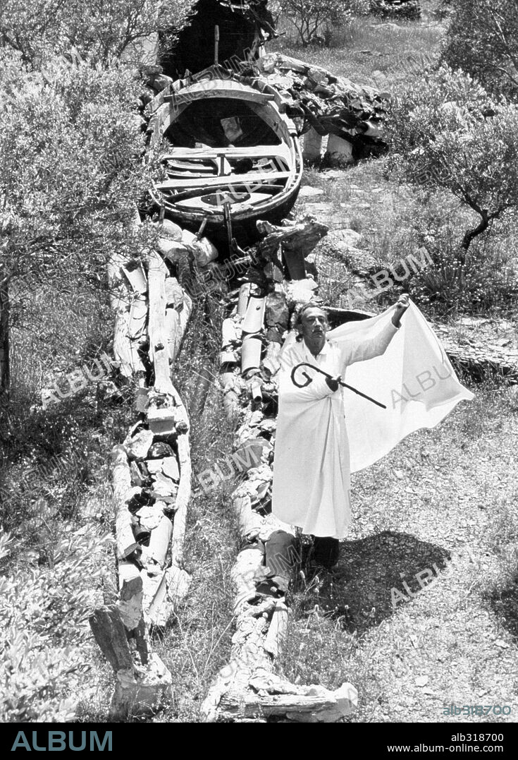 SALVADOR DALÍ. Salvador Dali, surrealist painted and sculptor, stands with his wife robe billowing him next to his giant figure of Christ which is constructed from Civil War debris.