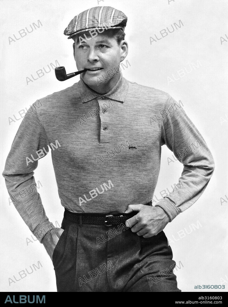 United States: c. 1954 A fashionable man smoking a pipe and wearing casual sporting attire of a Lacoste pullover shirt and a flat golf cap.