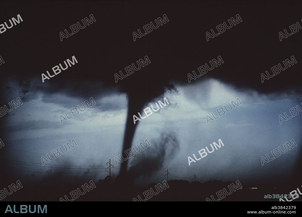 Tornado. Funnel cloud of a tornado seen between two electricity pylons. A tornado consists of a violently rotating column of air below a thundercloud. The tornado is visible because of the dust it picks up and the condensation of water droplets within it. Tornadoes can cause much destruction to property. The rotating air has a speed of 480 kilometers an hour or more. The width of the destructive path is usually a few hundred meters. The tornado moves along with its parent cloud, its path length may exceed 100 kilometers. Photographed in Kansas, USA, during spring. Tornadoes can occur at any time of the year, but the peak season of occurrence varies with the location.