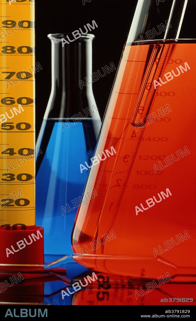 Laboratory glassware. Assortment of glassware containing colored solutions, used in chemistry research. At left is a measuring cylinder with yellow fluid; conical flasks are at center and right. Two pipettes are seen: one at lower frame, and one in the flask at right.