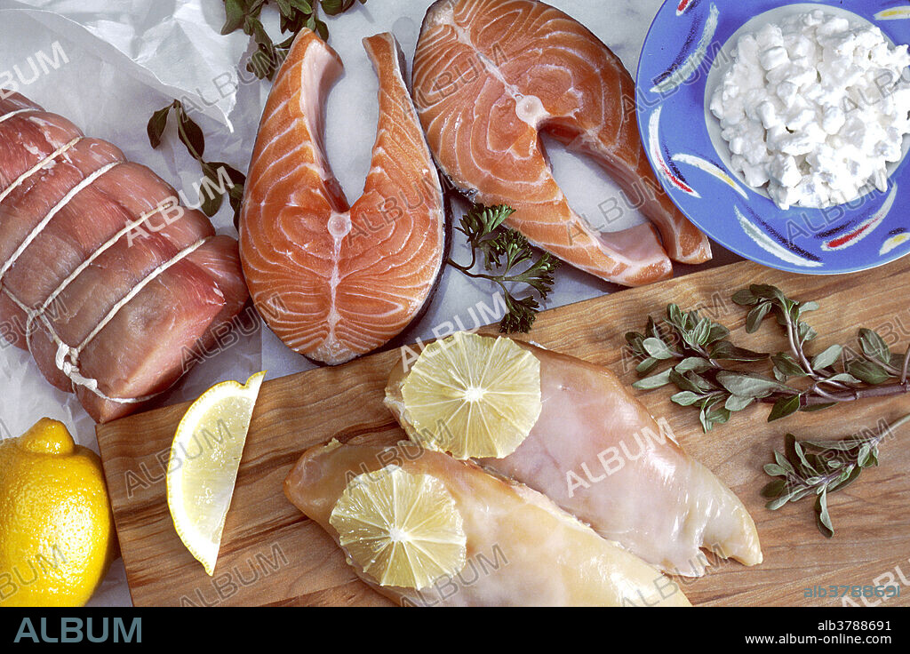 Wooden cutting board on a white tablecloth. There are various cuts of meat and fish on both the table and the block, garnished with sliced lemons, watercress and parsley. A blue bowl in the background is full of cottage cheese. Protein is a substance found in foods (such as meat, milk, eggs, and beans) that is an important part of the human diet.