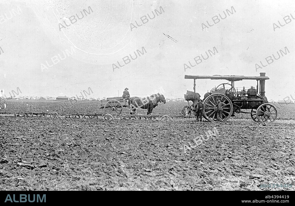 Negative - Steam Ploughing with Traction Engine Pulling Three Ploughs, Werribee, Victoria, 1910, Steam traction engine (Aveling & Porter?) pulling three ploughs with a horse and cart. There are two haystacks in the background. There are two men standing in front of the engine, they are wearing hats, trousers and jackets.
