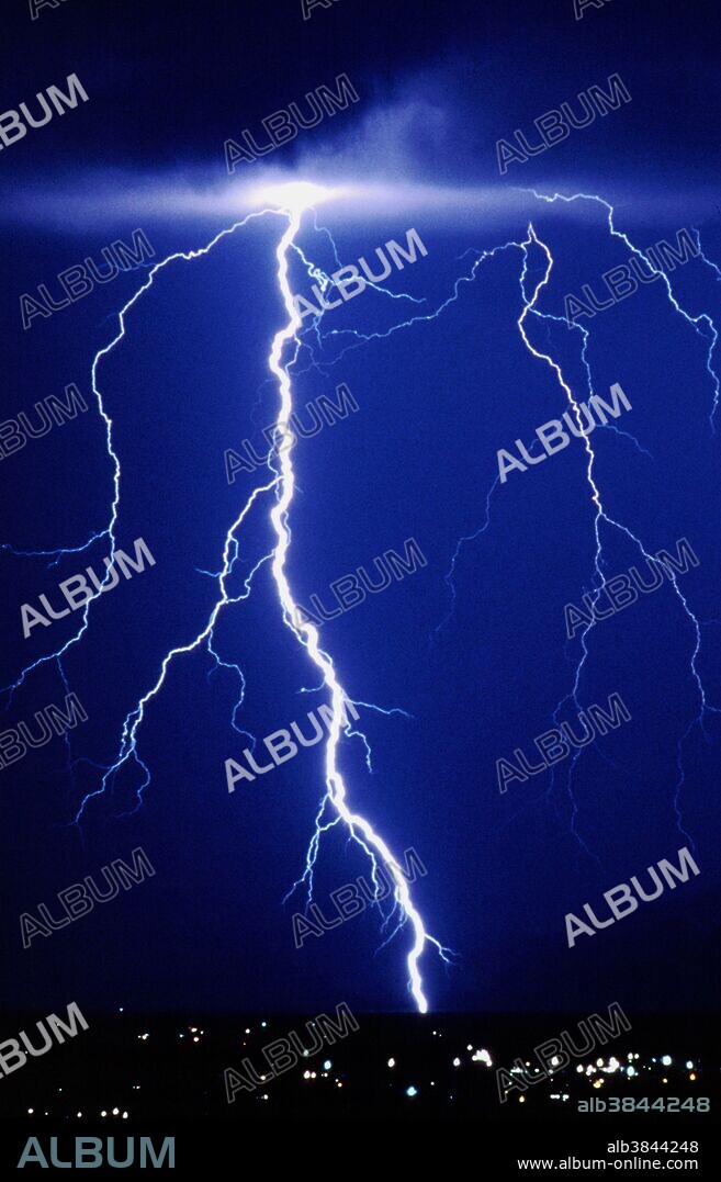 Lightning. Branching lightning bolt at night over a city. Lightning occurs when a large electrical charge builds up in a cloud; a potential difference of 10,000 volts a centimeter is needed for electrical discharge through air. At first, leaders of electrons descend towards the ground. As soon as one makes contact an enormous return stroke surges up the leader's ionized path, producing blinding white light and temperatures as high as 30,000 degrees Celsius. The other leaders also discharge themselves through the one that made contact with the ground, producing the branching effect seen here. Photographed on a summer night over Tucson in Arizona, USA.