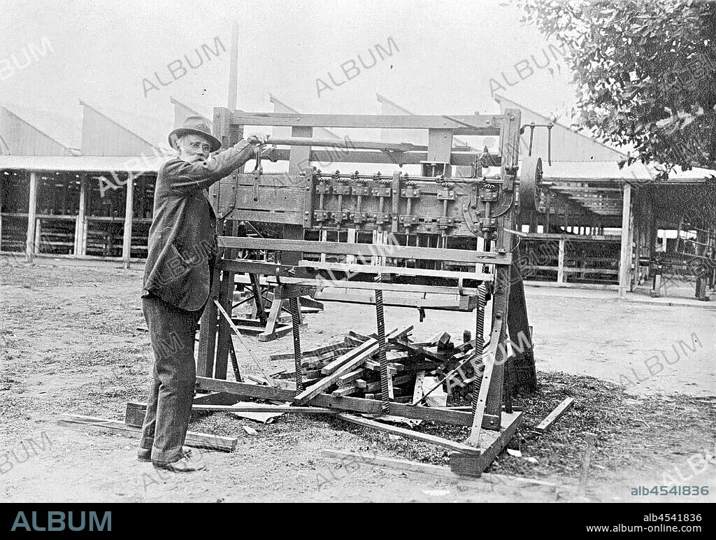 Negative - William Ross With Fence Dropper Boring Machine He Invented, Ballarat Showgrounds, Victoria, circa 1900, William Ross at the Ballarat Showgrounds with his invention of a dropper boring machine. He is wearing glasses, a hat and boots. There is a tree to the right of the image. A venue of the showgrounds is in the background.