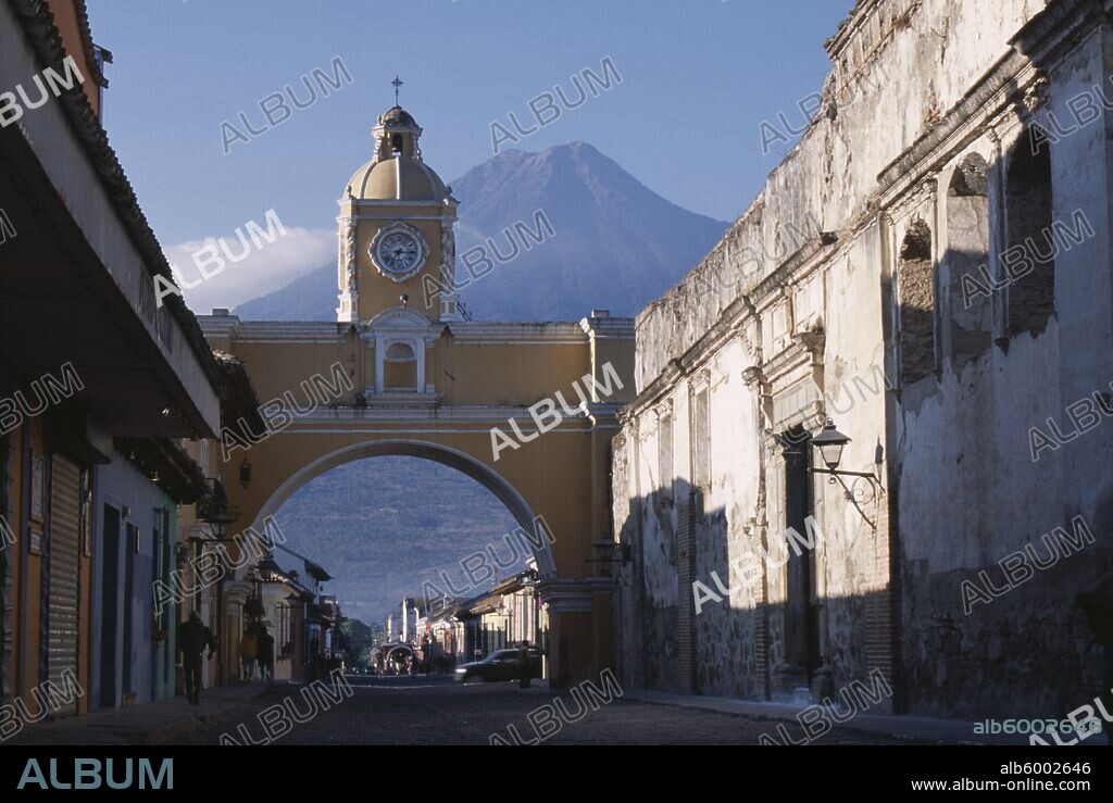 GUATEMALA Antigua Arco de Santa Catarina. Yellow painted archway with clock tower and people walking underneath with Volcan Agua beyond.