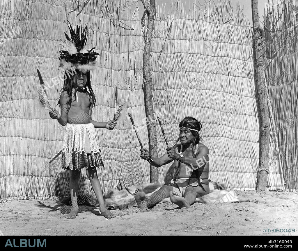 California: c. 1930 Medicine men taught their songs and dances to young boys chosen to carry on the traditions of the tribe. This Yokut medicine man is playing his split clap sticks by shaking them in time to his song. The dancing costume is a skirt of eagle down tipped with red-tailed hawk feathers. The hat is a basket decorated with eagle down, and raven and magpie feathers.
