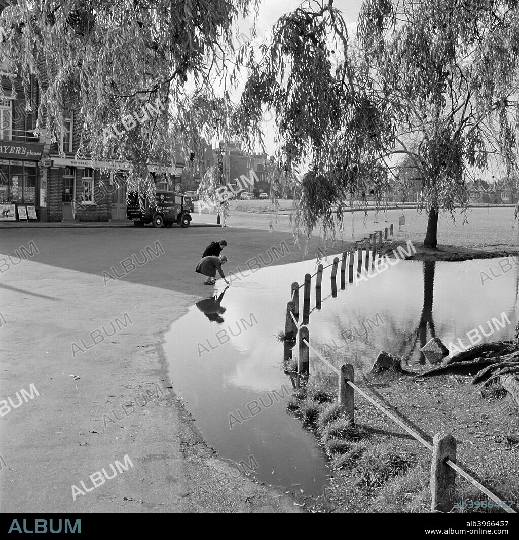 Hare and Billet Pond, Blackheath, London, 1955-1965. General view looking past the Hare and Billet public house on Grote's Place, towards Eliot Road and Hare and Billet Road. Two small girls are fishing with a net in the overflowing pond in the foreground.