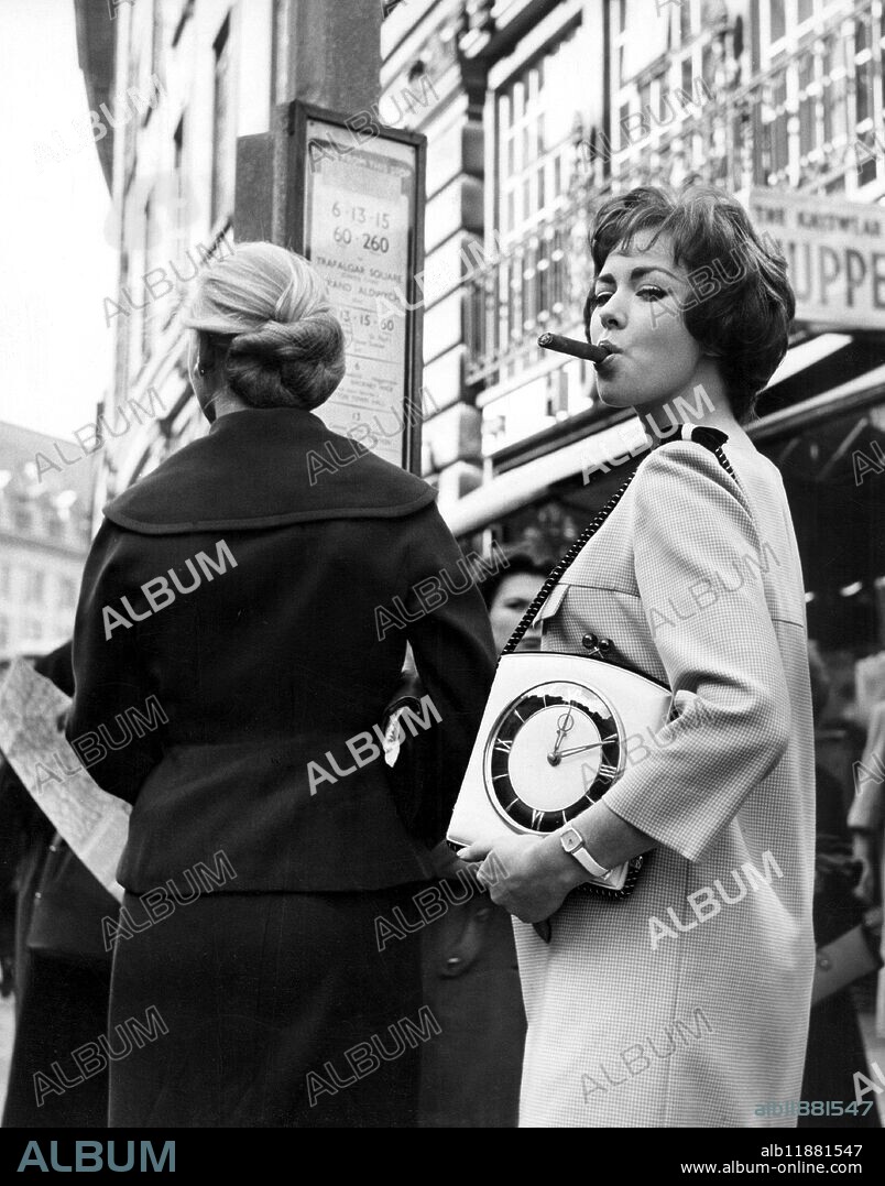 Beth Rogan at the height of fashion, smoking a cigar and holding a handbag with inlaid clock. c.1950s.