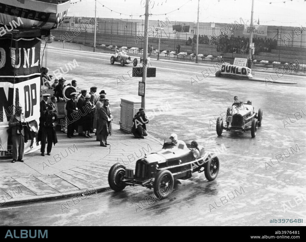 Brian Lewis in an Alfa Romeo Monza in the Mannin Moar race, Douglas, Isle of Man, 1933. Lewis won the race. Ahead of him is George Eyston also in an Alfa Romeo Monza. Eyston finished 3rd. Both Alfas are being pursued by a Bugatti. Dunlop banners can be seen.