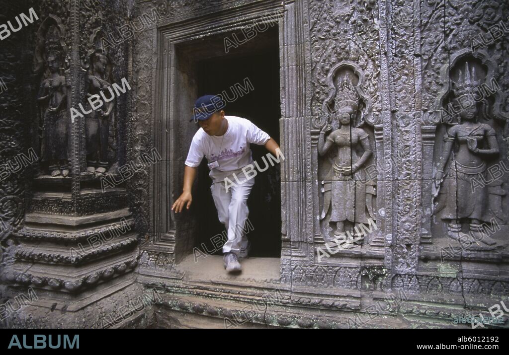 CAMBODIA Siem Reap Province Angkor Ta Prohm. Visitor climbing through doorway of twelth century Buddhist temple with exterior wall decorated with bas relief carvings.