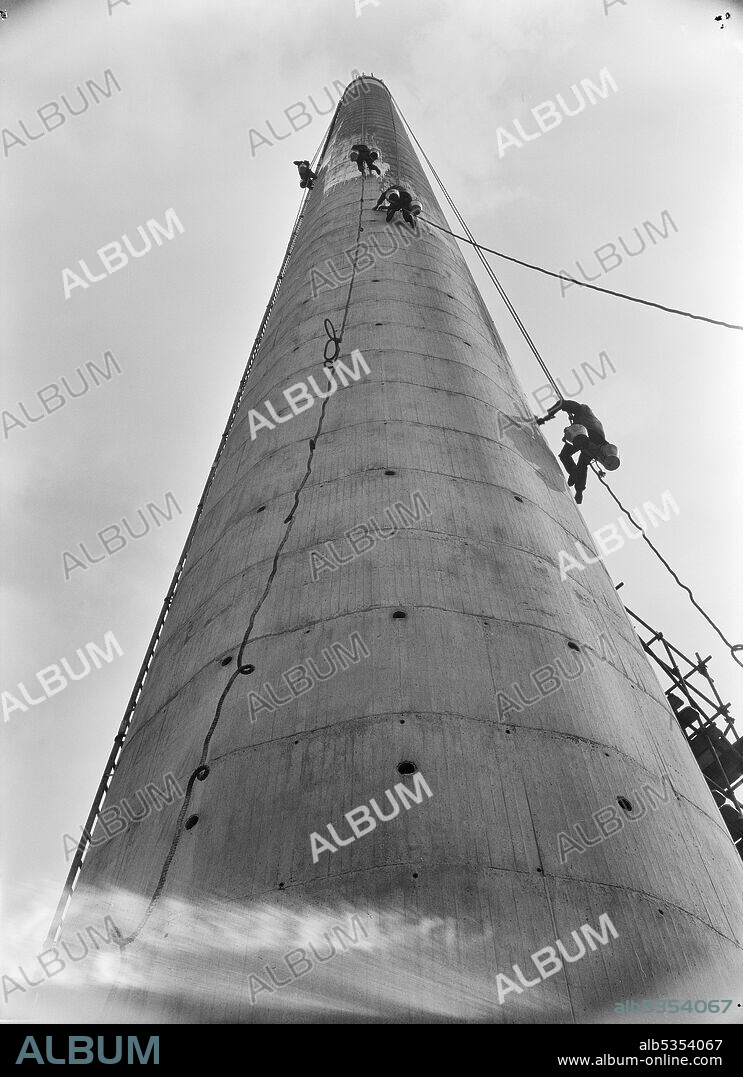 JOHN LAING PLC. Construction workers suspended in harnesses working on the surface of a chimney stack at the South Eastern Gas Board's (SEGAS) Power Station on the Isle of Grain. In the late 1950's, the South Eastern Gas Board (SEGAS) set up a power plant on the Isle of Grain. The plant was adjacent to the Grain refinery, and was designed to produce gas from petroleum products. In the negative register, 'Lytag S. E. Gas Board, Isle of Grain, Kent' is recorded next to this image. Lytag is a lightweight aggregate material that can withstand high temperatures. According to the April 1963 edition of Laing's monthly newsletter, Team Spirit, Lytag was used for the flue lining at the SEGAS Isle of Grain plant.