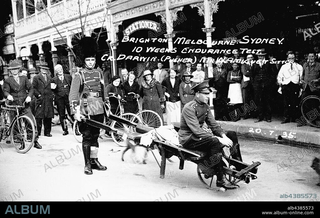 Negative - Wangaratta, Victoria, 1935, A man in military uniform pushing another on a wheelbarrow as part of an endurance test from Melbourne to Sydney. The uniforms do not appear to be regular service uniforms. The Bull's Head and Albion hotels are in the background.