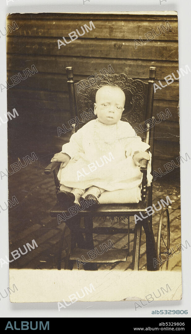 A photographic postcard of an infant seated on a wooden stroller. The baby is wearing a gown and booties and is holding onto the stroller's arm rests and sitting on folded cloth. The back of the photographic postcard is unused and has [POST CARD] printed in black at the top and blank spaces for [CORRESPONDENCE] and [ADDRESS]. In the upper right corner there is a [AZO] stamp.