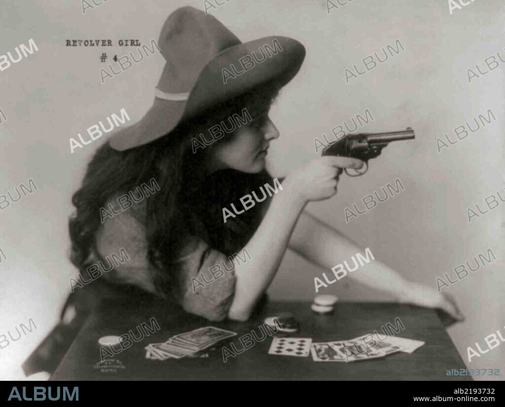 Woman in cowgirl clothing, seated at table with deck of cards and chips, pointing pistol, with arm on table.