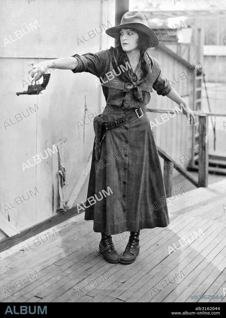 Hollywood, California:  c. 1919. Famous Players Lasky actress Edna Mae Cooper gingerly holds a pistol by its trigger guard. She says she did not feel comfortable in Western togs.