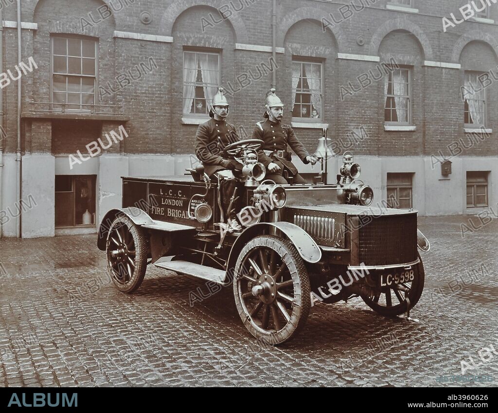 Firemen in brass helmets aboard a motor hose tender, London Fire Brigade Headquarters, London, 1909. Moustached firemen pose on a vehicle; one is ringing the bell.