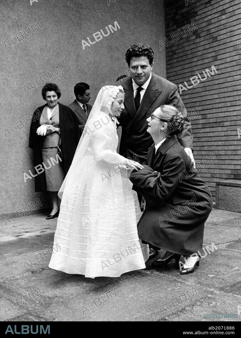 Luigi Infantino and Sarah Ferrati at the holy communion of their daughter. Italian actress Sarah Ferrati crouching at the feet of her daughter Monica Infantino, looking into her eyes. Behind them, her husband, Italian tenor Luigi Infantino. 1961.
