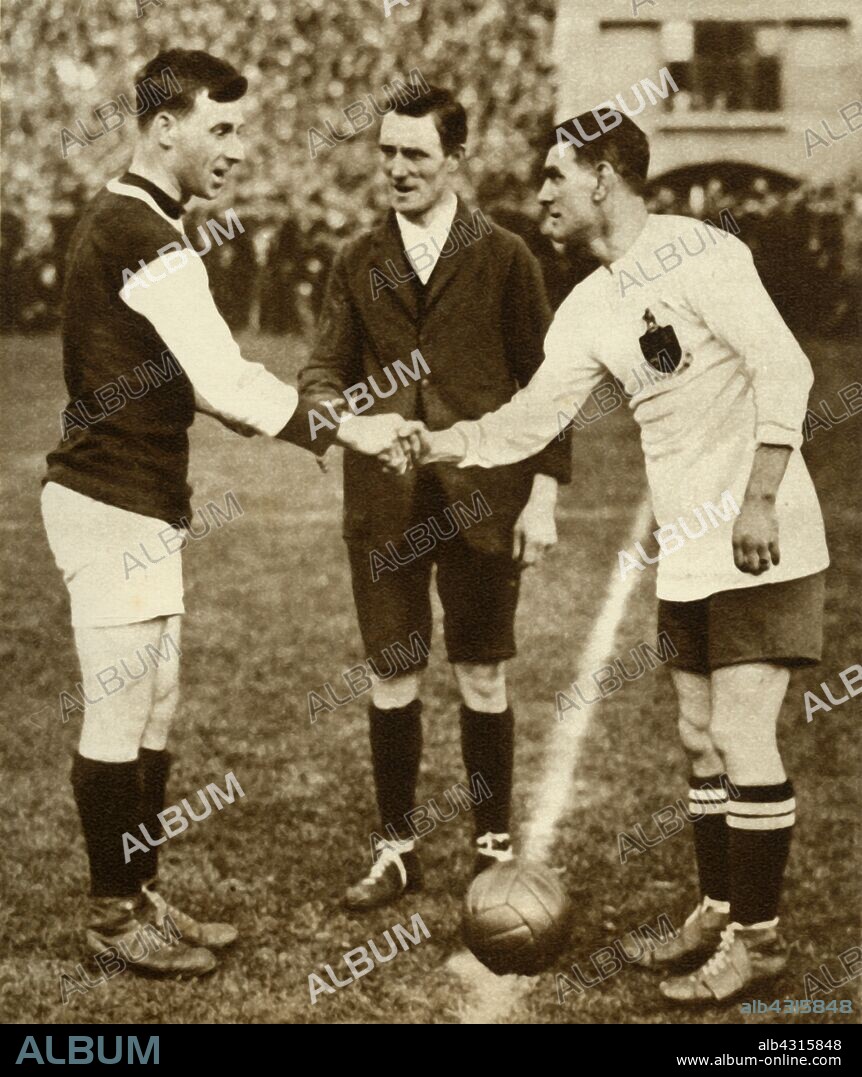 George Kay and Joe Smith shake hands before kick-off, FA Cup Final, Wembley Stadium, London, 28 April 1923, (1935). George Kay (1891-1954), captain of West Ham United, shaking hands with Joe Smith (1889-1971), the Bolton Wanderers' captain, as the first FA (Football Association Challenge Cup) Final is held at the new Wembley Stadium. Bolton Wanderers beat West Ham 2-0. From "The Silver Jubilee Book - The Story of 25 Eventful Years in Pictures". [Odhams Press Ltd., London, 1935].