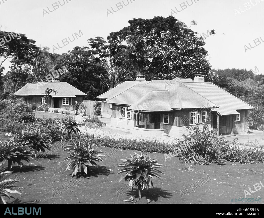 Kenya, Rear elevation of Sagana Lodge, a ground floor timber building on a stone basement with a tiled roof set amongst trees and copious flower boundaries. In the foreground is a garden with unidentified plants growing in it. A secondary, smaller building is set back from the main lodge, February 1952. 2001/090/1/4/1/70.