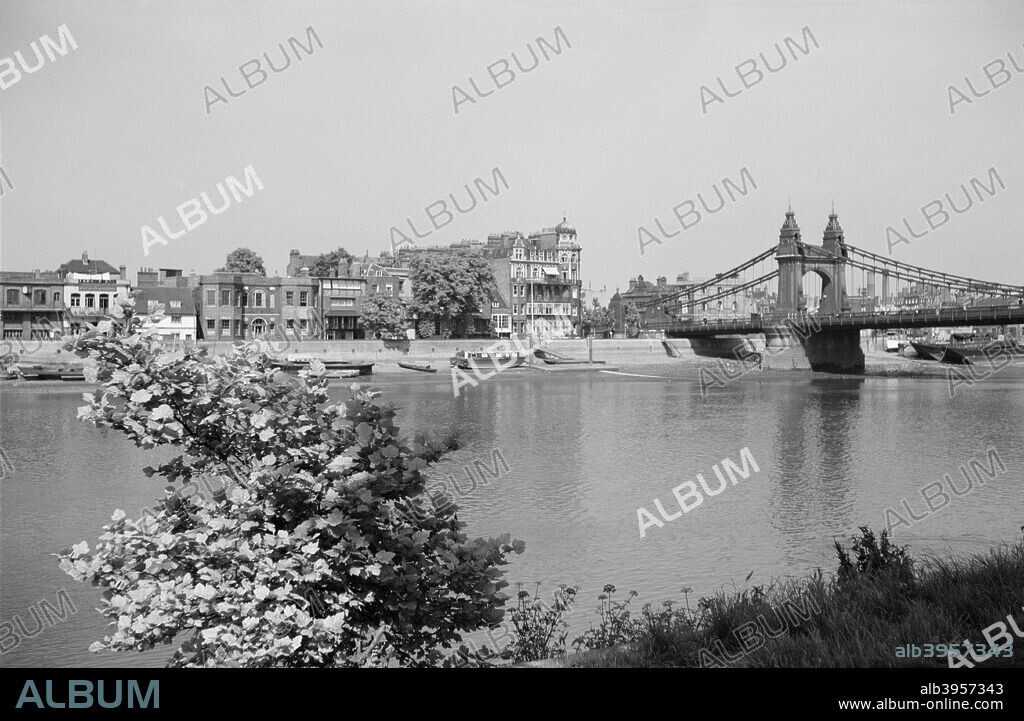 River Thames, Hammersmith, London, c1945-1965. The waterfront at Hammersmith viewed from the opposite shore with part of Hammersmith Suspension Bridge, designed by Sir Joseph Bazalgette and opened in 1887, on the right.