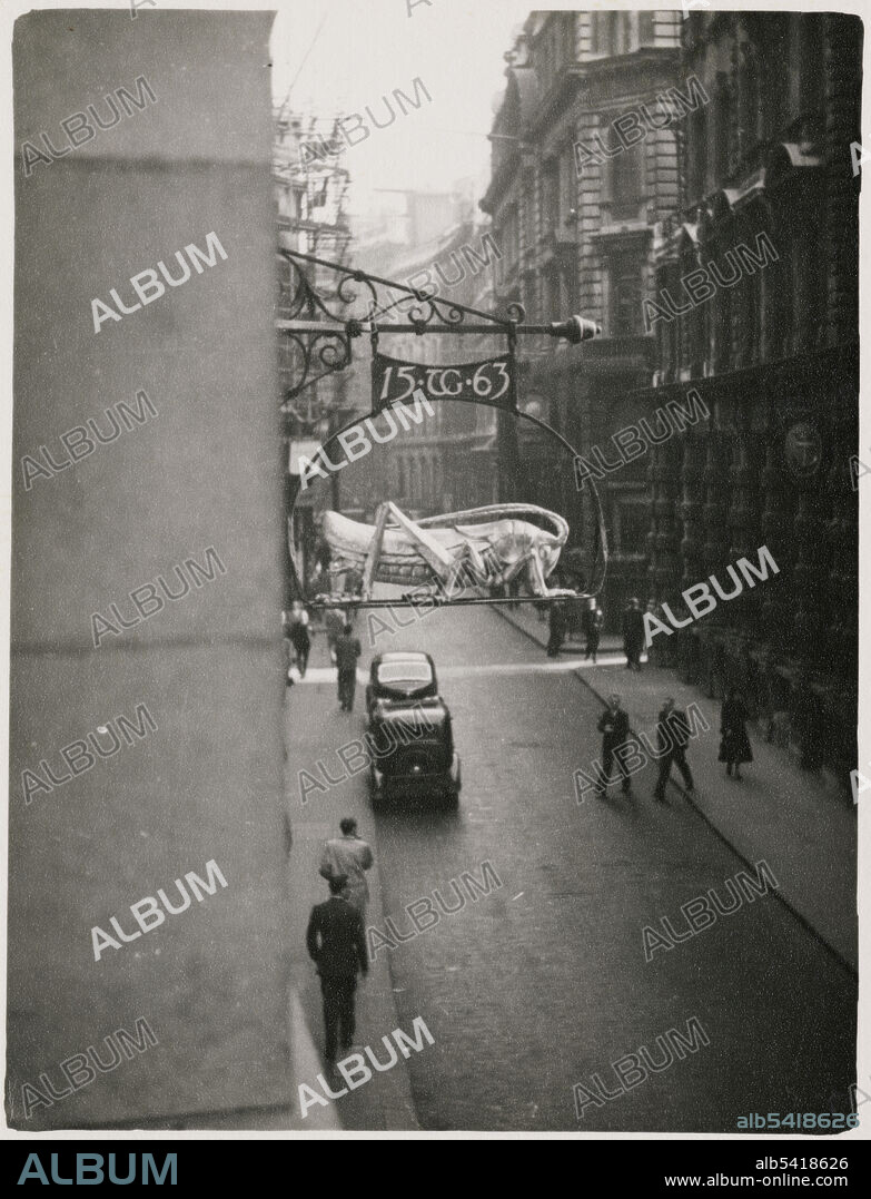 JR UPPINGTON. Martins Bank, Lombard Street, City and County of the City of London, Greater London Authority, 1950-1953. Looking east along Great George Street towards Big Ben and Parliament Square, with pedestrians on the pavement in the foreground. The sign of the grasshopper was the crest of the Gresham family, and was used to distinguish the house of Thomas Gresham in Lombard Street in the 16th century. Gresham was a financier to Edward VI, Mary I and Elizabeth I, and founder of the Royal Exchange. The sign bears Gresham's initials and the date 1563; it is believed he traded at the site from this date. The freehold of the Grasshopper, later numbered 68 Lombard Street, was purchased by John Martin in the 1740s. Martins Bank was demolished and rebuilt in 1930. The bank remained at the site until it merged with Barclays in 1969; the grasshopper was still hung outside 68 Lombard Street into the 21st century. It is believed that 68 Lombard Street is the longest continuous use of business premises as a bank.