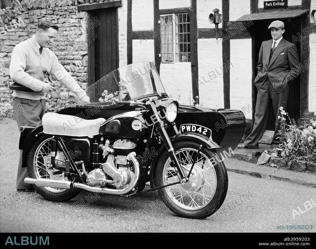 An Ariel VB 600cc with a sidecar, 1956. Parked outside a cottage, with a smiling woman sitting in the sidecar. The company was founded by James Starley and William Hillman in 1870 to make bicycles and the first patented tensioned wire-spoke wheel. Ariel first made a powered tricycle in 1898 using a de Dion engine. They made their first motorized two-wheeler in 1901, powered by Kerry engines, selling them in 1902. The company closed in 1970.