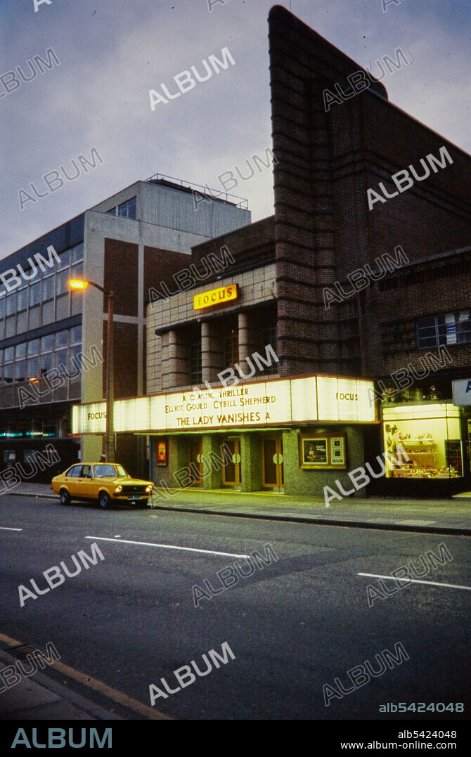NORMAN WALLEY. Focus Cinema, Delamere Street, Crewe, Cheshire East, 1975-1983. An exterior view of the Focus Cinema at dusk, showing the illuminated billboard. The building was designed as the Odeon Cinema by Budge Reid of Harry W Weedon's practice, and was opened in July 1937. The cinema was taken over by Brent Walker in October 1975 and was renamed the Focus Cinema. It was closed in May 1983 and was demolished in September 1983.