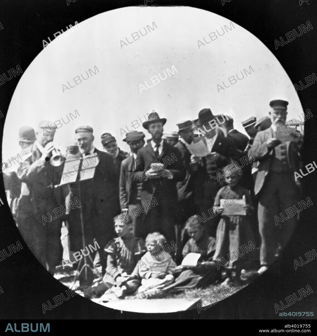 London City Mission (LCM) group at Barnet Fair, London, 1900s. Men singing hymns while a man accompanies them on a trumpet. A group of gypsy children are in the foreground. Galt came to London in 1890 to work as a missionary for the London City Mission. In the early 1900s he took a series of photographs, mainly of the East End, which he had made into lantern slides to illustrate lectures publicising the work of the Mission. Galt sought to show that though there was great poverty in the East End, the people were not sub-human, as was popularly imagined by the middle-classes, but ordinary folk trying to do their best under difficult circumstances.