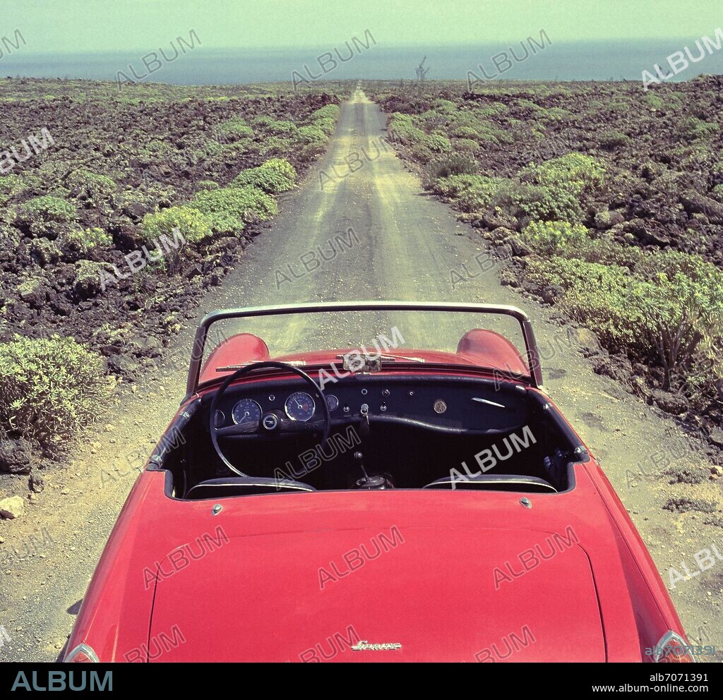 AUSTIN HEALEY SPRITE MK III DESCAPOTABLE ROJO AÑOS 60 EN UN CAMINO RECTO ENTRE LA LAVA.