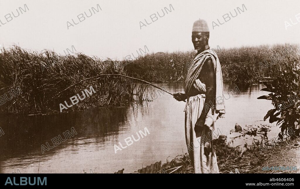 Sudan, Printed caption reads: 'Fishing on the Jur River, Bahr-El-Ghazal. Published by G N Morhig, The English Pharmacy, Khartoum. Copyright 61'. A man with a rod stands on the bank of a river, [c.1906]. 2003/222/1/2/59.