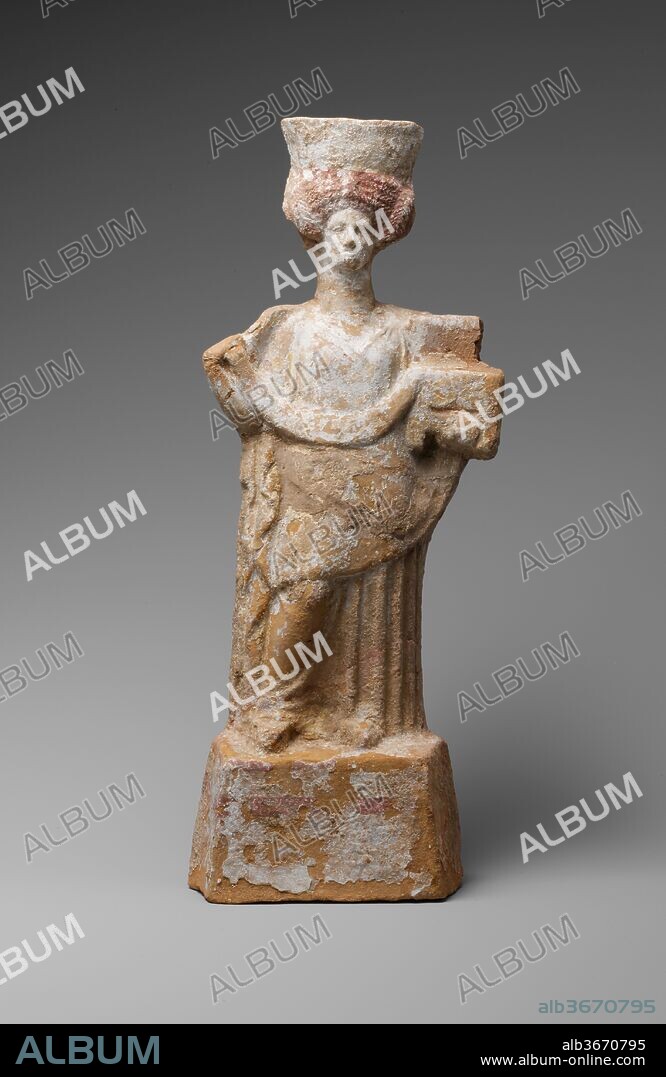 Terracotta statuette of a standing woman with a basket and wreath. Culture: Greek, Boeotian. Dimensions: h. 14 1/8 in. (35.9 cm). Date: late 5th century B.C..
Weight on left leg, right knee bent. Left arm supports an open casket with legs, from which a fillet is drawn across the waist by the raised right hand (right hand may never have been fully rendered). Peplos is open down right side; hem of knee-length overfold is drawn up toward left. Head type is characteristic of this group - long wide neck with small face, elaborate wig. This wig has four rows of braid above forehead, with coiled plaits over ears. High, wide polos. High, splaying, trapezoidal base.