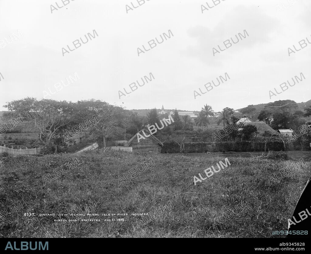 Distant View Womens Prison, Isle of Pines, Noumea, Burton Brothers studio, photography studio, 1899, Dunedin, black-and-white photography, From rise above settlement, with thatched roofs and wooden and thatched fence around. Tall tropical trees including coconut trees and in distance on hill above colonial buildings including a church with spire.